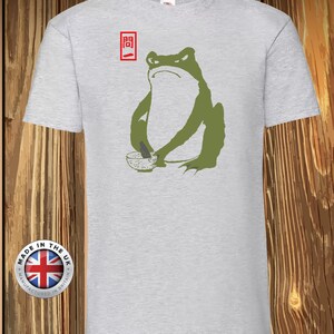 Könnte beinhalten: Ein hellgraues Kurzarm-T-Shirt mit einer Grafik eines grünen Frosches, der eine kleine Schale hält. Über dem Frosch befindet sich ein rotes Rechteck mit den japanischen Schriftzeichen 問一. Das T-Shirt hat einen klassischen Rundhalsausschnitt.