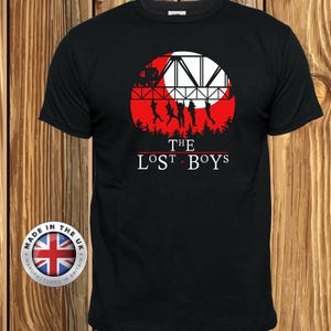 May include: Black t-shirt featuring a red and white graphic of a bridge, silhouettes of figures, and the text "THE LOST BOYS." The design is set within a red circle. A circular emblem with the Union Jack flag is on the lower left.