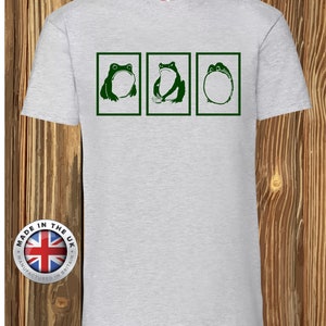 Op de afbeelding: Een lichtgrijs T-shirt met korte mouwen en drie donkergroene rechthoekige kaders. Elk kader bevat een andere illustratie van een kikker: één zittend, één met een klein kommetje en één die zich tot een ronde vorm heeft opgerold.