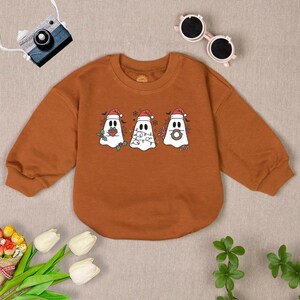 Puede incluir: Una sudadera marr&oacute;n de manga larga con cuello redondo presenta un gr&aacute;fico de tres fantasmas con gorros de Pap&aacute; Noel. Los fantasmas sostienen un regalo, una cadena de luces y una corona.