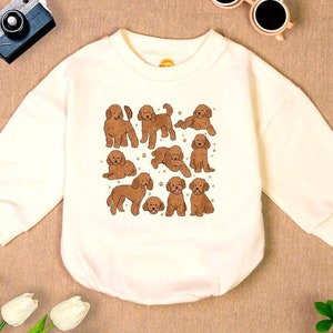 May include: A white long-sleeved sweatshirt with a crew neck and a graphic print of brown poodles in various poses. The poodles are surrounded by small white paw prints.
