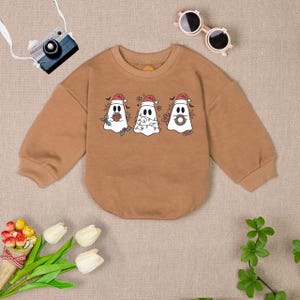 Puede incluir: Una sudadera marr&oacute;n de manga larga con un dise&ntilde;o de fantasma navide&ntilde;o. Los fantasmas llevan gorros de Pap&aacute; Noel y tienen decoraciones festivas como regalos, luces y coronas.