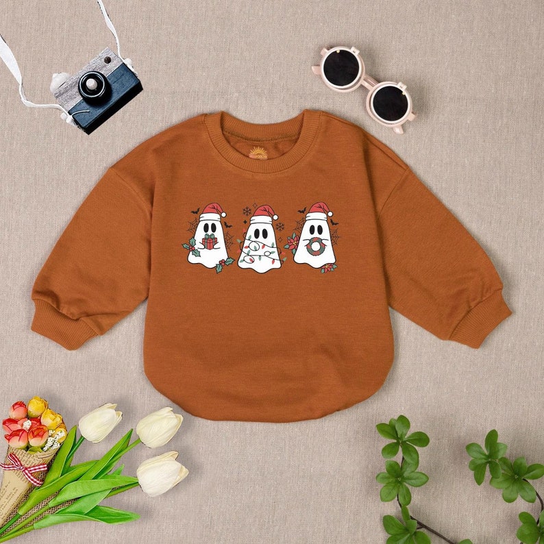Puede incluir: Una sudadera marr&oacute;n de manga larga con cuello redondo presenta un gr&aacute;fico de tres fantasmas con gorros de Pap&aacute; Noel. Los fantasmas sostienen un regalo, una cadena de luces y una corona.