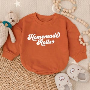 May include: An orange baby onesie with the words "Homemade Rolls" in white script. The onesie has long sleeves and a round neckline. The image also includes a stuffed bear, baby shoes, and wooden beads.