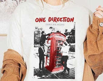 May include: White t-shirt featuring a graphic of a red telephone booth with figures, and the text "ONE DIRECTION TAKE ME HOME" in red. The design is a black and white image with a pop of red.