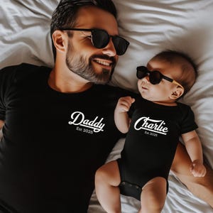 May include: A black t-shirt with the word "Daddy" and a black baby onesie with the name "Charlie", both with "Est 2025" in white script. Both wear sunglasses. The image is taken from above.