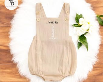 May include: Tan baby romper with Amelia printed above a candle graphic and the word "one". The romper has button closures on the straps and is displayed on a white fluffy surface with a bouquet of white flowers.