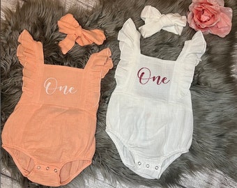 May include: Two white baby onesies with ruffled sleeves. One onesie is peach and the other is white. Both onesies have the word "One" embroidered on the front. The peach onesie has a white bow headband and the white onesie has a white headband.