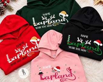 May include: Four hooded sweatshirts in dark green, red, black, and light pink. Each features a graphic with the text "NAME", "Lapland", and "YOUR TEXT HERE 2026". The designs include festive elements like reindeer antlers, elf legs, and Santa hats.
