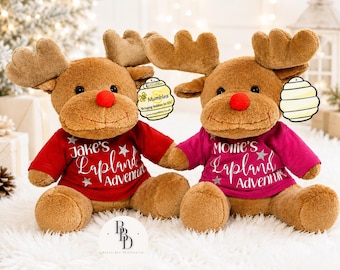 May include: Two plush reindeer toys with brown fur and red noses. The left reindeer wears a red shirt with white text reading Jake's Lapland Adventure and silver stars. The right reindeer wears a magenta shirt with white text reading Mollie's Lapland Adventure and silver stars.