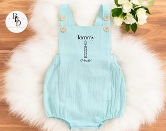 May include: A light blue baby romper with wooden button closures. The romper has the name "Tommy" and a candle graphic with the word "one" printed on the front. The romper is displayed on a white fluffy surface.