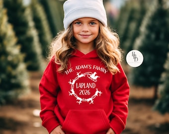 May include: A bright red hooded sweatshirt featuring a white graphic print of a reindeer-pulled sleigh in a circular pattern. The text reads THE ADAM'S FAMILY, LAPLAND, and 2026. The hoodie includes a front kangaroo pocket and long sleeves.