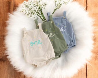 May include: Three corduroy baby rompers in beige, green, and blue, each with the word "one" or "me" embroidered on the front. The rompers have buttoned straps and are arranged on a white fluffy surface with white flowers.