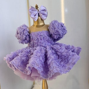 Pu&ograve; includere: Un abito color lavanda con un designo a rose 3D. L'abito ha maniche a sbuffo e una gonna ampia. Un fiocco lavanda con dettagli di perle &egrave; sulla parte superiore dell'abito.