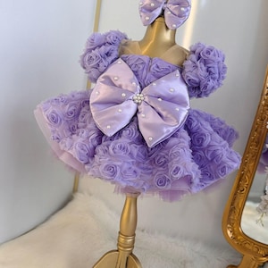 Pu&ograve; includere: Un abito color lavanda con un grande fiocco di raso e decorazioni di perle. L'abito &egrave; realizzato in tulle e presenta un design a rosa 3D. L'abito &egrave; su un supporto dorato.