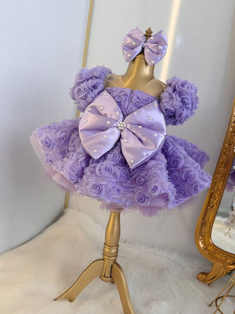 Pu&ograve; includere: Un abito color lavanda con un grande fiocco di raso e decorazioni di perle. L'abito &egrave; realizzato in tulle e presenta un design a rosa 3D. L'abito &egrave; su un supporto dorato.