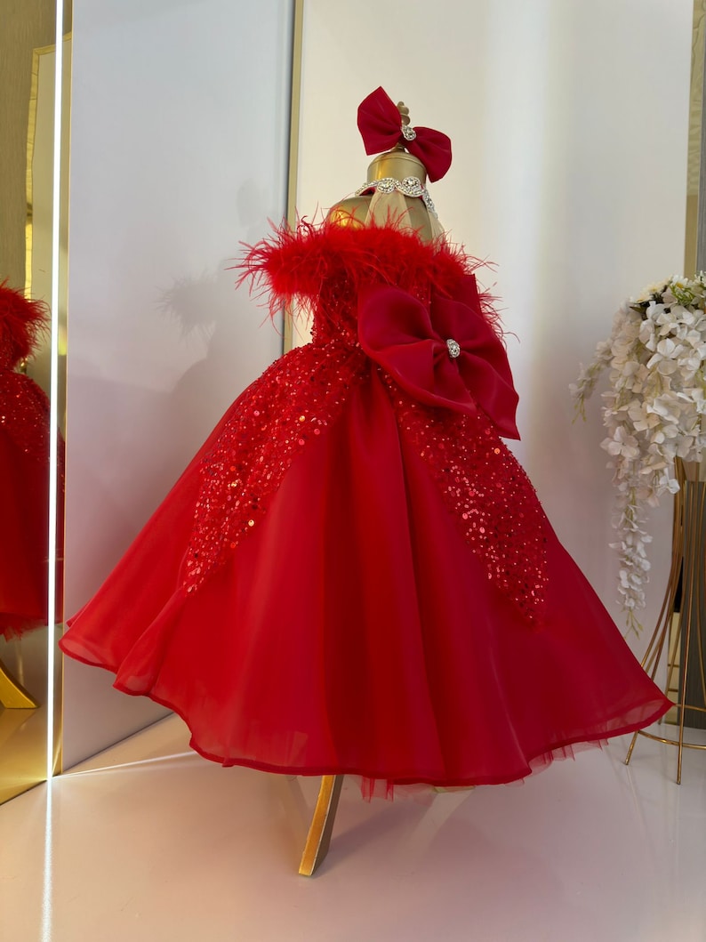 Puede incluir: Un vestido de tul rojo con un gran lazo rojo y ribete de plumas. El vestido est&aacute; decorado con lentejuelas y tiene una falda completa.