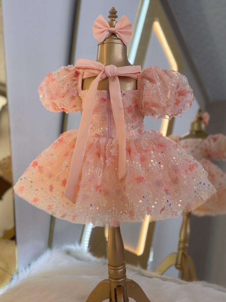 May include: A pink tulle dress with a floral pattern and sequins. The dress has puffed sleeves and a bow at the back. The dress is on a gold mannequin.
