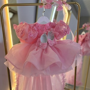 Puede incluir: Un vestido rosa para beb&eacute; con detalles de mariposas, una diadema a juego y zapatos. El vestido tiene una falda en capas y se exhibe en una percha dorada. El conjunto est&aacute; dise&ntilde;ado para una ocasi&oacute;n especial.