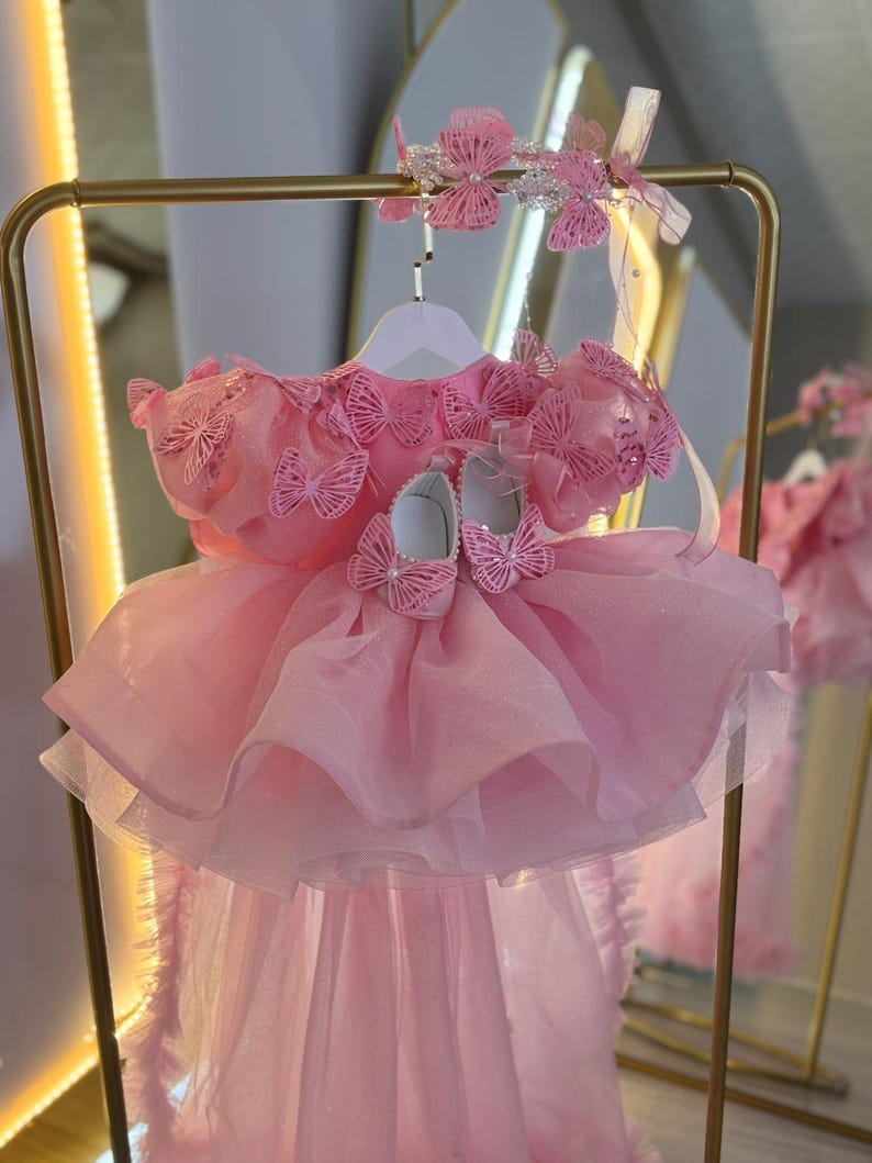 Puede incluir: Un vestido rosa para beb&eacute; con detalles de mariposas, una diadema a juego y zapatos. El vestido tiene una falda en capas y se exhibe en una percha dorada. El conjunto est&aacute; dise&ntilde;ado para una ocasi&oacute;n especial.