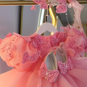 Puede incluir: Un vestido rosa con detalles de mariposas, una diadema a juego y un par de zapatos. El vestido presenta una falda en capas y est&aacute; adornado con delicados recortes de mariposas. Los zapatos tienen detalles de perlas.