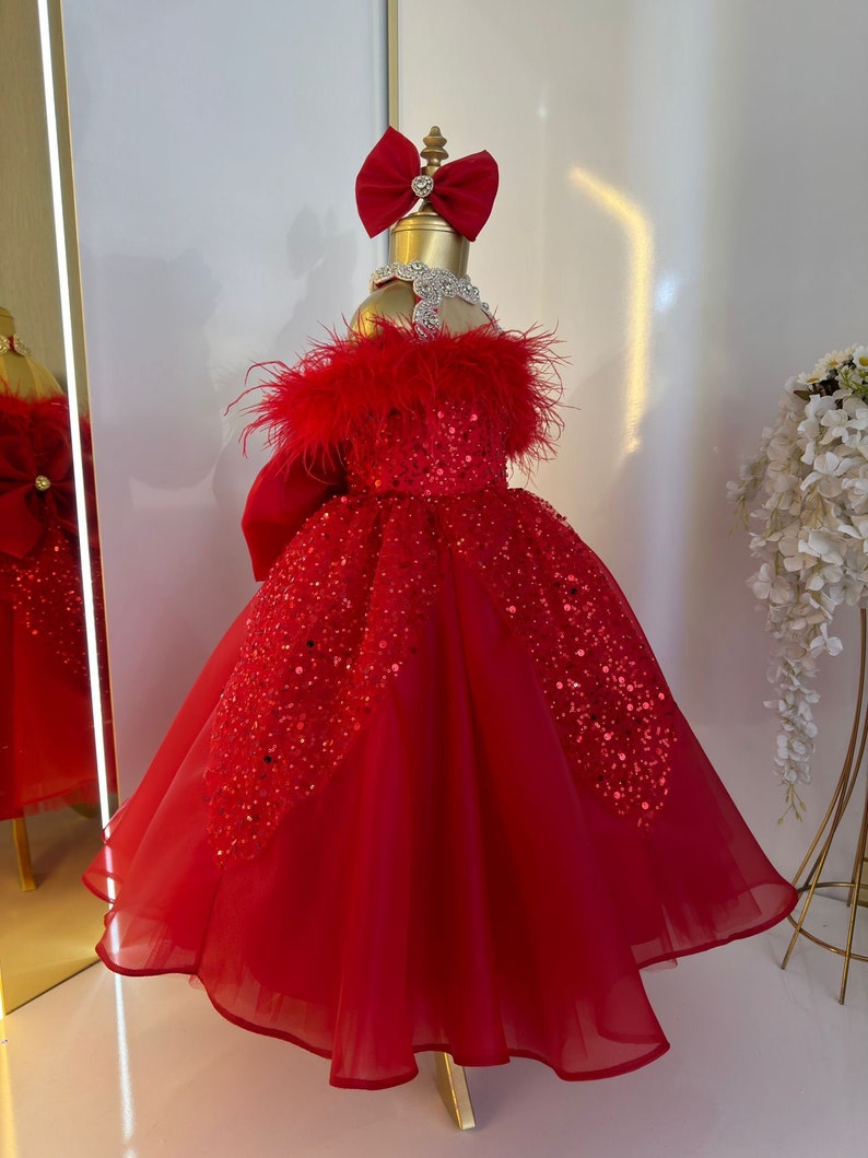 Puede incluir: Un vestido de tul rojo con lentejuelas y ribete de plumas. El vestido tiene un lazo en la parte superior y est&aacute; en un maniqu&iacute;.