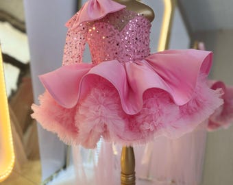 May include: A pink, one-shoulder dress with a sequined bodice and a layered skirt. The dress is displayed on a gold mannequin and features a matching headband with a bow. The skirt has a ruffled, fluffy texture.