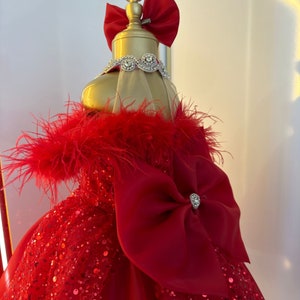Puede incluir: Un vestido rojo de lentejuelas con un gran lazo rojo y ribete de plumas en los hombros. El vestido est&aacute; en un maniqu&iacute; con un lazo rojo en la cabeza.