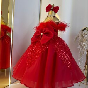 Puede incluir: Un vestido de tul rojo con un gran lazo rojo y detalles de lentejuelas. El vestido tiene un escote de plumas y un soporte dorado.