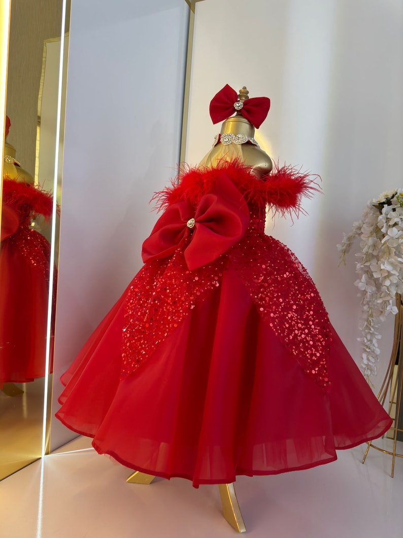 Puede incluir: Un vestido de tul rojo con un gran lazo rojo y detalles de lentejuelas. El vestido tiene un escote de plumas y un soporte dorado.