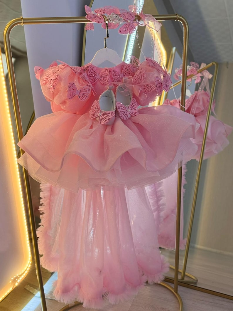 Puede incluir: Un vestido de beb&eacute; rosa con una falda en capas y detalles de mariposas. El vestido se exhibe con zapatos y una diadema a juego, todos adornados con delicados detalles de mariposas. El vestido est&aacute; colgado de una percha blanca.