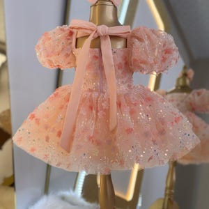 May include: A pink tulle dress with a floral pattern and sequins. The dress has puffed sleeves and a bow at the back. The dress is on a gold mannequin.