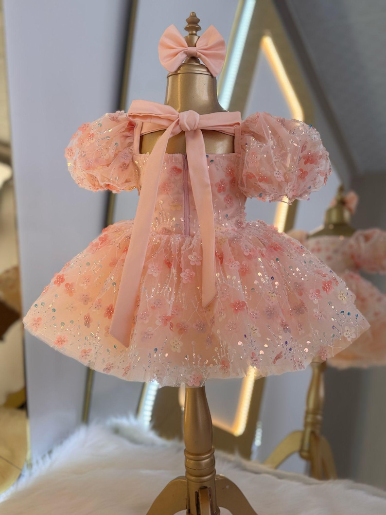 May include: A pink tulle dress with a floral pattern and sequins. The dress has puffed sleeves and a bow at the back. The dress is on a gold mannequin.