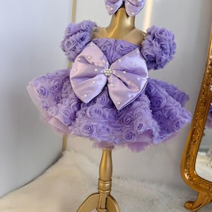 Pu&ograve; includere: Un abito color lavanda con un grande fiocco di raso e decorazioni di perle. L'abito &egrave; in tulle e ha una gonna a balze. L'abito &egrave; su un supporto dorato.