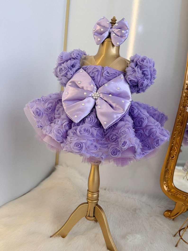 Pu&ograve; includere: Un abito color lavanda con un grande fiocco di raso e decorazioni di perle. L'abito &egrave; in tulle e ha una gonna a balze. L'abito &egrave; su un supporto dorato.