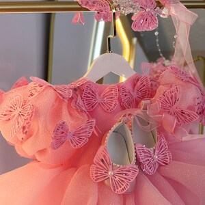 Puede incluir: Un vestido rosa con detalles de mariposas, una diadema a juego y zapatos. El vestido presenta m&uacute;ltiples adornos de mariposas y una falda de tul. Los zapatos est&aacute;n adornados con perlas y detalles de mariposas. La diadema tiene detalles de mariposas y cuentas.