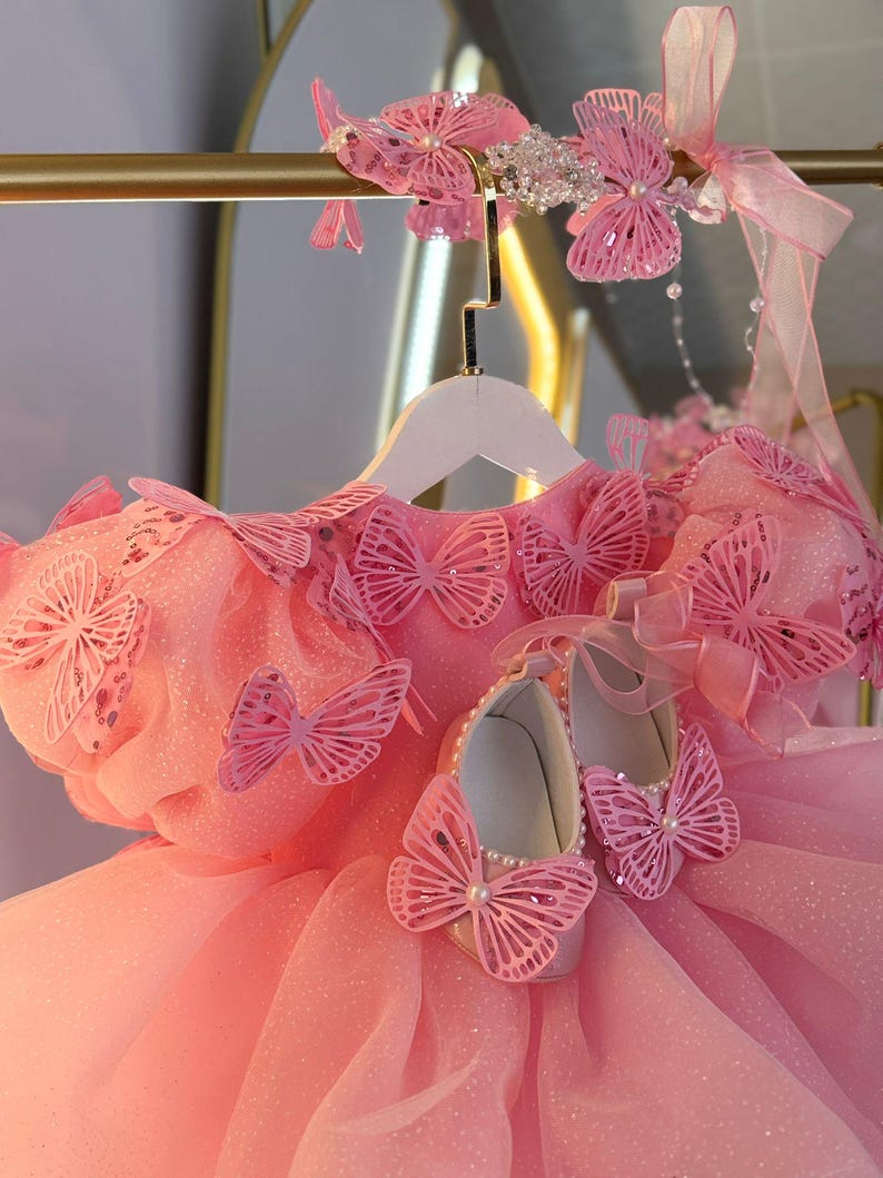 Puede incluir: Un vestido rosa con detalles de mariposas, una diadema a juego y zapatos. El vestido presenta m&uacute;ltiples adornos de mariposas y una falda de tul. Los zapatos est&aacute;n adornados con perlas y detalles de mariposas. La diadema tiene detalles de mariposas y cuentas.