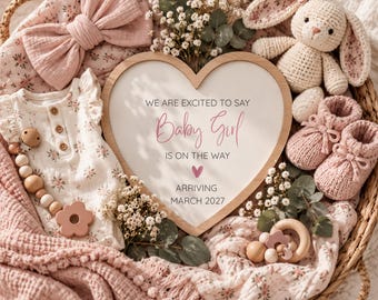 May include: A wicker basket containing a heart-shaped sign that reads WE ARE EXCITED TO SAY Baby Girl IS ON THE WAY ARRIVING MARCH 2027, a floral onesie, a pink bow headband, a crochet bunny, pink knit booties, a wooden pacifier clip, and baby blankets.