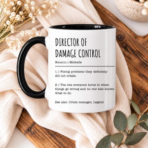 May include: A white ceramic mug with a black handle and interior. The mug features black text that reads: DIRECTOR OF DAMAGE CONTROL, Noun(n.) Michelle, 1.) Fixing problems they definitely did not create. 2.) The one everyone turns to when things go wrong and no one else knows what to do. See also: Crisis manager, Legend.