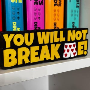 May include: A black and yellow sign with the text "YOU WILL NOT BREAK ME!" and a pair of heart-patterned underwear. The sign is on a white shelf with colorful books in the background.
