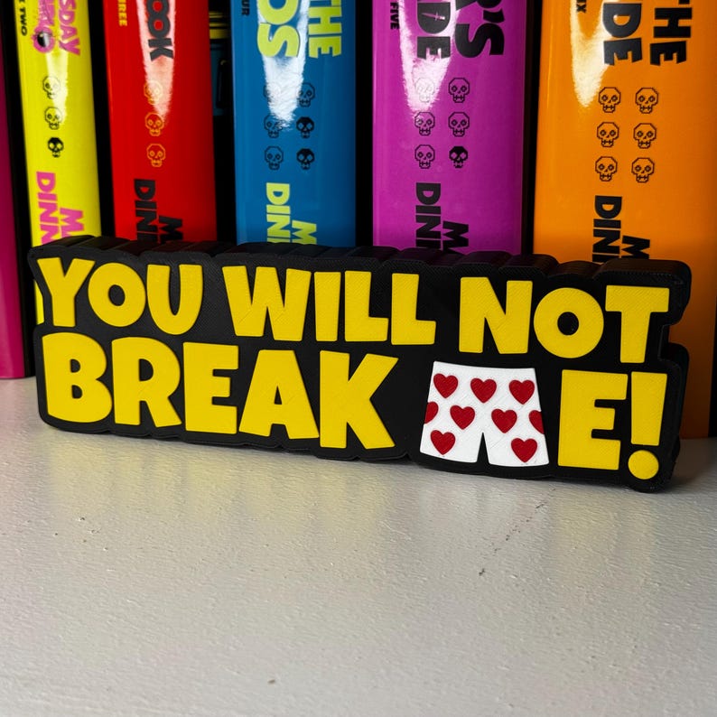 Może przedstawiać: Czarny prostokątny znak z ż&oacute;łtym napisem YOU WILL NOT BREAK E! Litera A w słowie BREAK została zastąpiona grafiką białych bokserek z powtarzającym się wzorem małych czerwonych serc na przedniej powierzchni.