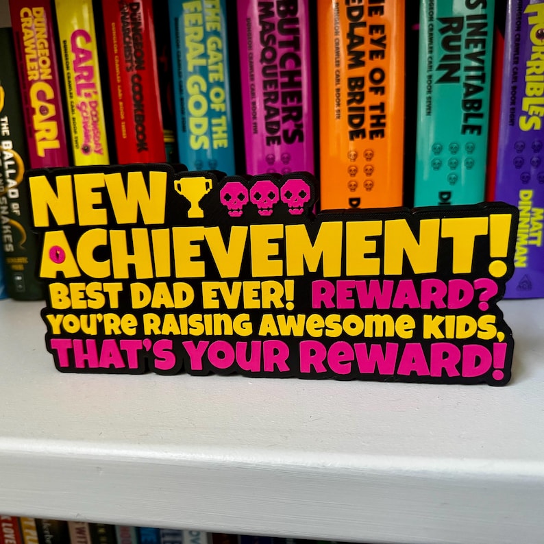 Może przedstawiać: Prostokątny znak z czarnym tłem, zawierający ż&oacute;łty i r&oacute;żowy tekst: NEW ACHIEVEMENT! BEST DAD EVER! REWARD? YOU'RE RAISING AWESOME KIDS, THAT'S YOUR REWARD! Znak zawiera ż&oacute;łtą ikonę trofeum oraz trzy r&oacute;żowe czaszki.
