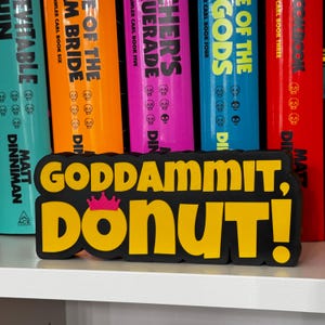 Może przedstawiać: Czarno-ż&oacute;łty znak wydrukowany w technologii 3D z napisem "GODDAMMIT, DONUT!" pogrubioną czcionką bezszeryfową. Mała, jaskrawor&oacute;żowa ikona korony znajduje się nad literą O w słowie DONUT. Znak posiada grubą czarną obw&oacute;dkę.