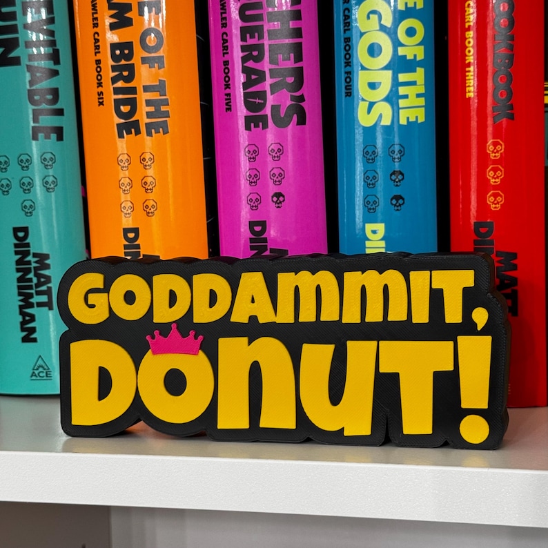 Może przedstawiać: Czarno-ż&oacute;łty znak wydrukowany w technologii 3D z napisem "GODDAMMIT, DONUT!" pogrubioną czcionką bezszeryfową. Mała, jaskrawor&oacute;żowa ikona korony znajduje się nad literą O w słowie DONUT. Znak posiada grubą czarną obw&oacute;dkę.