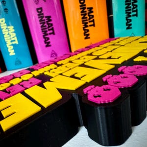 Może przedstawiać: Czarny znak wydrukowany w technologii 3D z wypukłymi ż&oacute;łtymi i r&oacute;żowymi literami oraz r&oacute;żowymi ikonami czaszek. W tle widoczne są cztery grzbiety książek, z kt&oacute;rych każdy zawiera nazwisko autora Matt Dinniman czarnym drukiem na niebieskiej, r&oacute;żowej, pomarańczowej i turkusowej okładce.