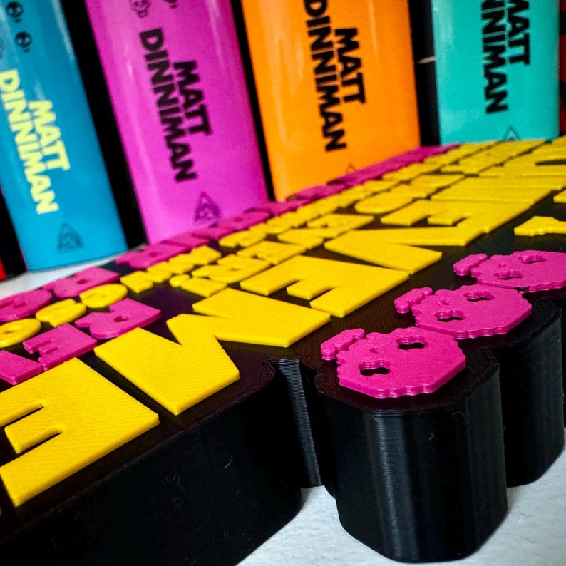 Może przedstawiać: Czarny znak wydrukowany w technologii 3D z wypukłymi ż&oacute;łtymi i r&oacute;żowymi literami oraz r&oacute;żowymi ikonami czaszek. W tle widoczne są cztery grzbiety książek, z kt&oacute;rych każdy zawiera nazwisko autora Matt Dinniman czarnym drukiem na niebieskiej, r&oacute;żowej, pomarańczowej i turkusowej okładce.