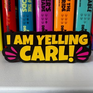 Może przedstawiać: Czarna prostokątna naklejka z ż&oacute;łtym napisem I AM YELLING CARL! Naklejka posiada stylizowane r&oacute;żowe akcenty po lewej i prawej stronie, przypominające linie ruchu lub fale dźwiękowe, umieszczona na tle kolorowych grzbiet&oacute;w książek.