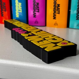 Może przedstawiać: Czarny, tr&oacute;jwymiarowy znak z wypukłym ż&oacute;łtym napisem "DUNGEON CRAWLERS" oraz małymi r&oacute;żowymi i ż&oacute;łtymi ikonami graficznymi. Znak znajduje się przed kilkoma kolorowymi grzbietami książek autorstwa Matta Dinnimana.