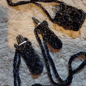 Może przedstawiać: Czarne i szare szydełkowe uchwyty na długopisy vape z pasującymi smyczami. Uchwyty wykonane są z miękkiego, pluszowego materiału. Jeden uchwyt jest przymocowany do małej torebki. Pi&oacute;ra vape są widoczne wewnątrz uchwyt&oacute;w.