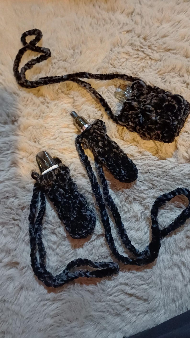 Może przedstawiać: Czarne i szare szydełkowe uchwyty na długopisy vape z pasującymi smyczami. Uchwyty wykonane są z miękkiego, pluszowego materiału. Jeden uchwyt jest przymocowany do małej torebki. Pi&oacute;ra vape są widoczne wewnątrz uchwyt&oacute;w.